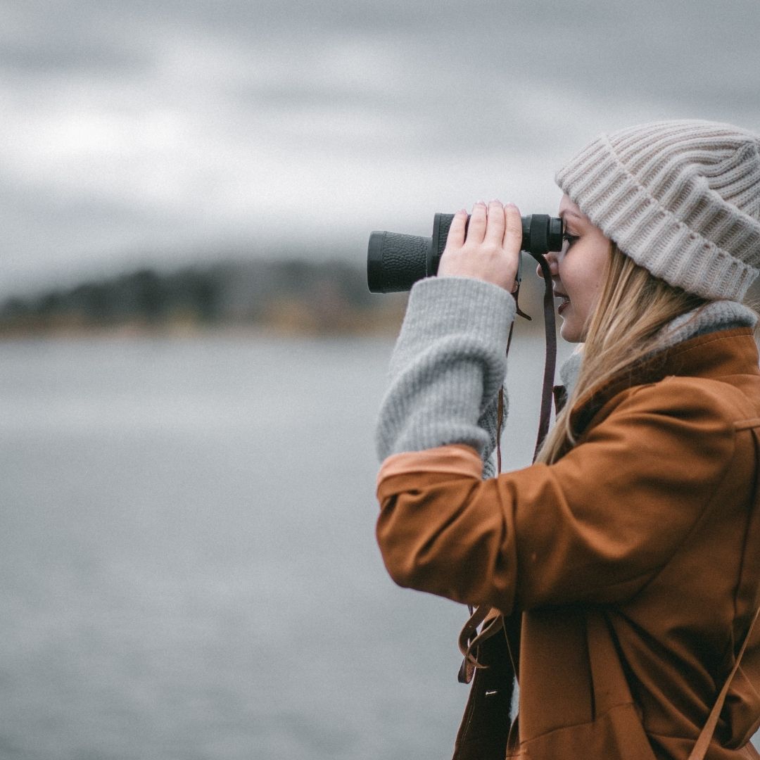Frau mit Mütze schaut mit Fernglas über das Wasser – achtsames Beobachten