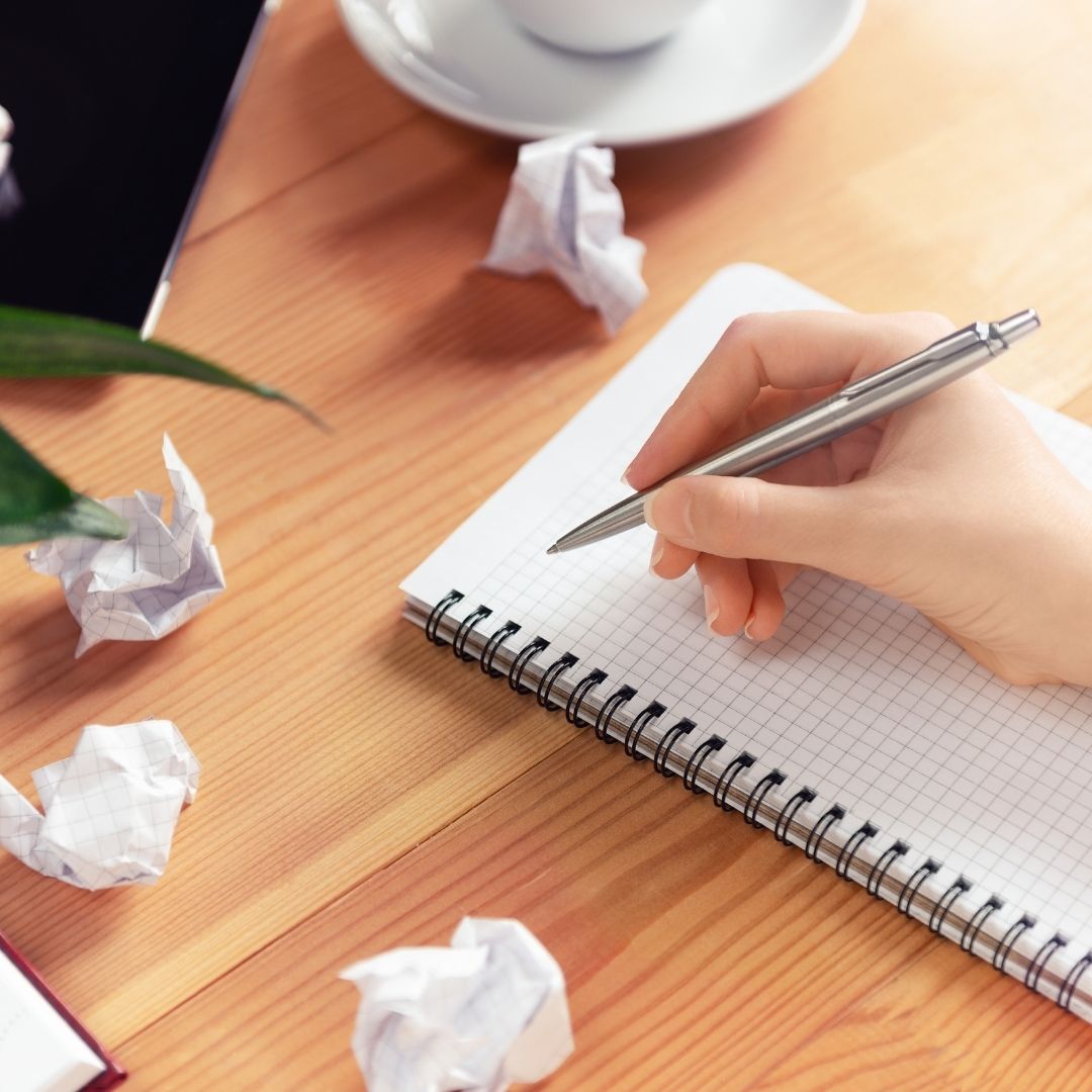 Hand schreibt in Notizbuch, zerknüllte Zettel daneben – beschreiben statt bewerten (nicht urteilen)