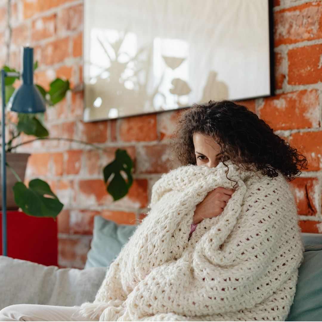 Person sitzt eingekuschelt unter einer Decke auf dem Sofa und nimmt sich fünf Minuten bewusste Pause